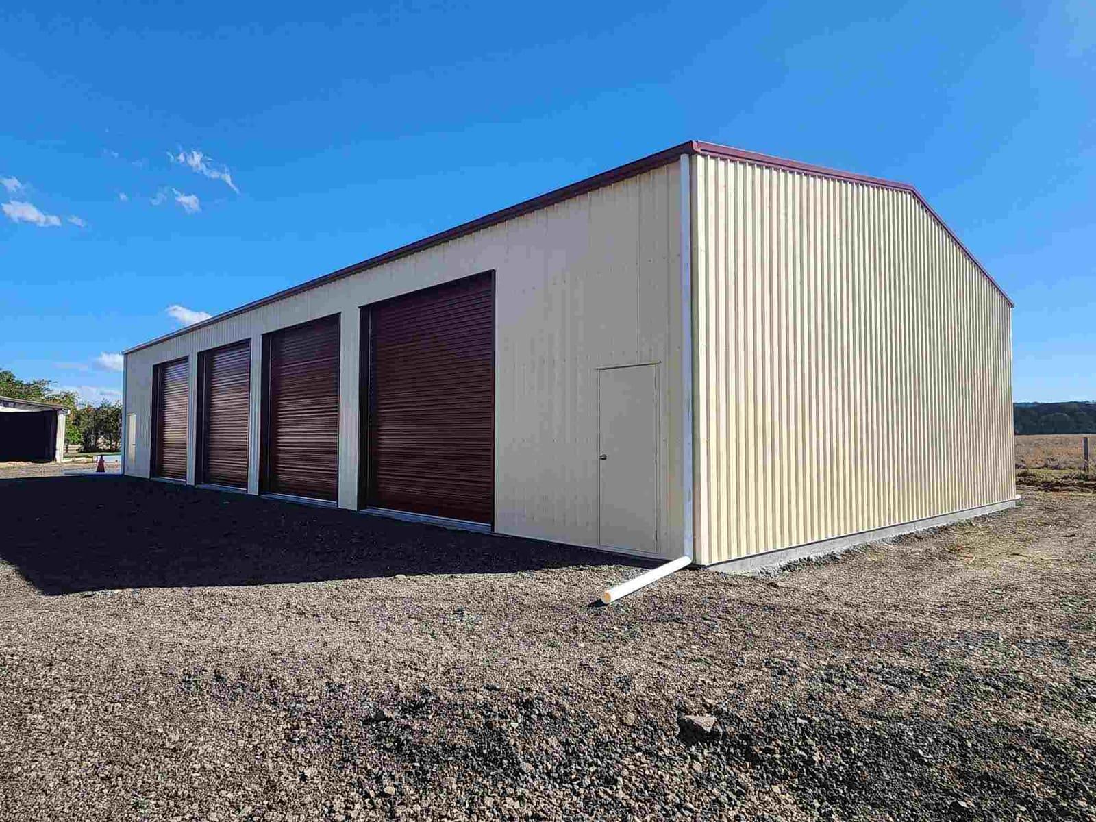 Cream industrial shed with red trim and four maroon roller doors on fresh gravel