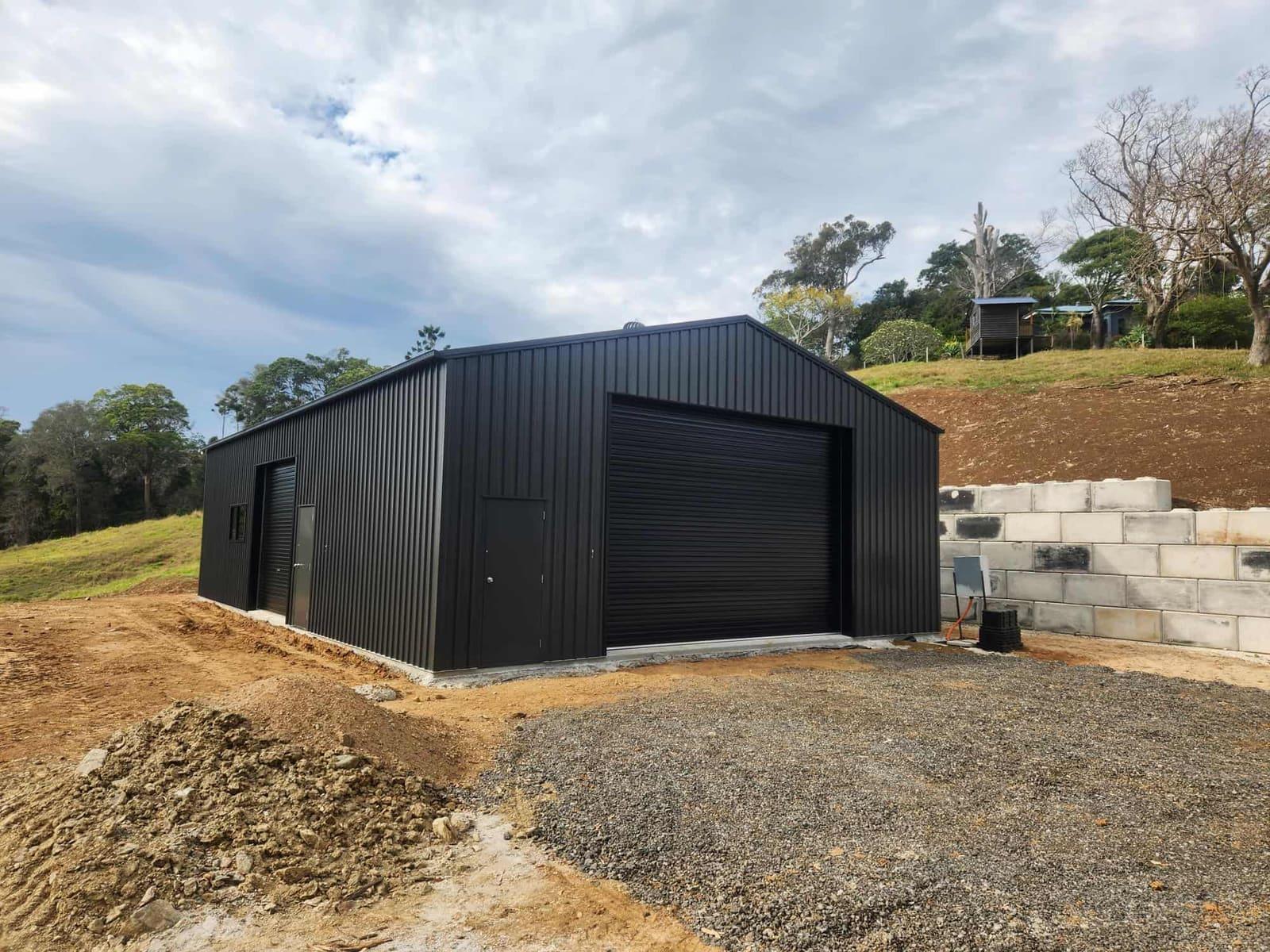 Black steel shed with a large roller door, block retaining wall and gravel apron